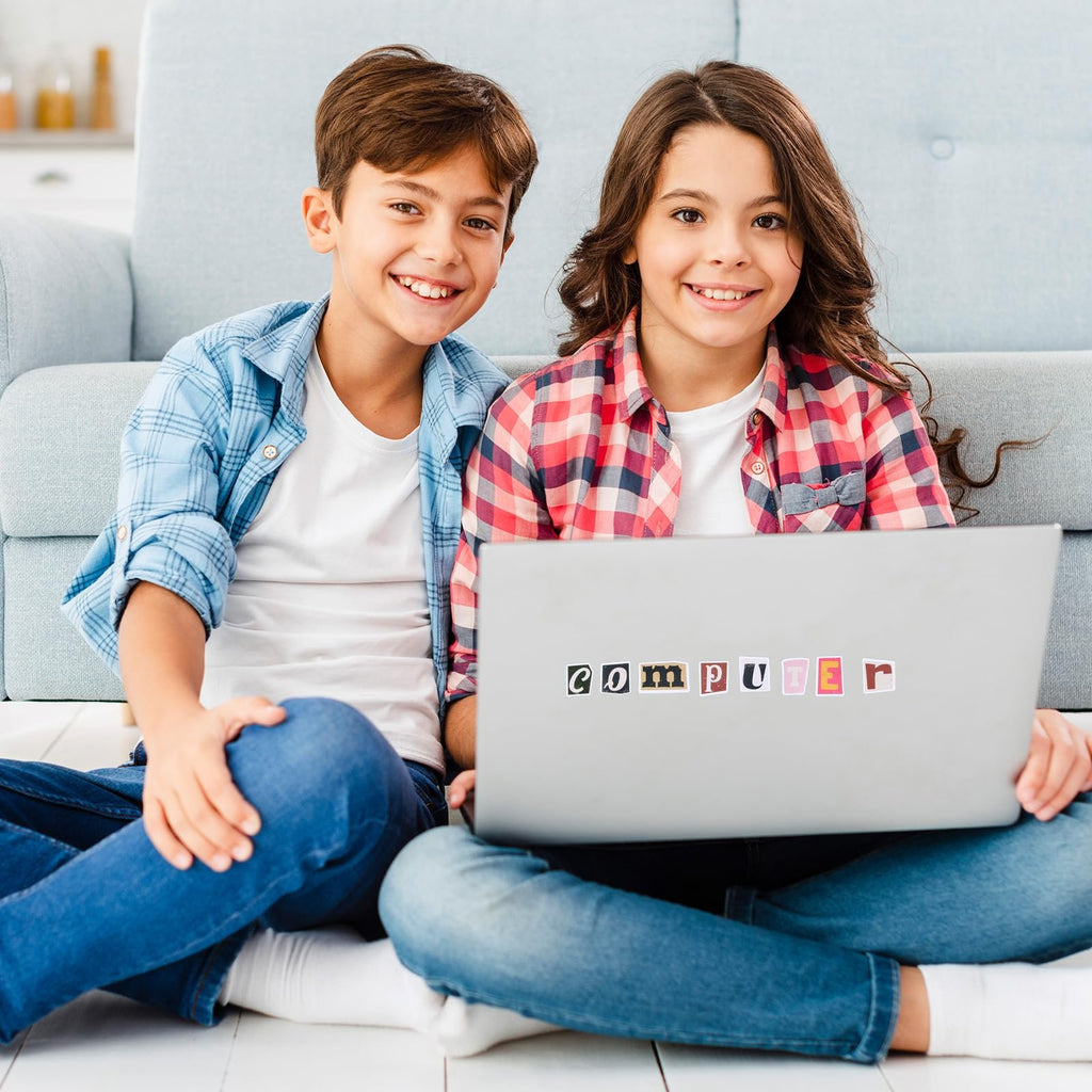 Two smiling children sitting cross-legged on the floor with a laptop, laptop has letter stickers spelling COMPUTER