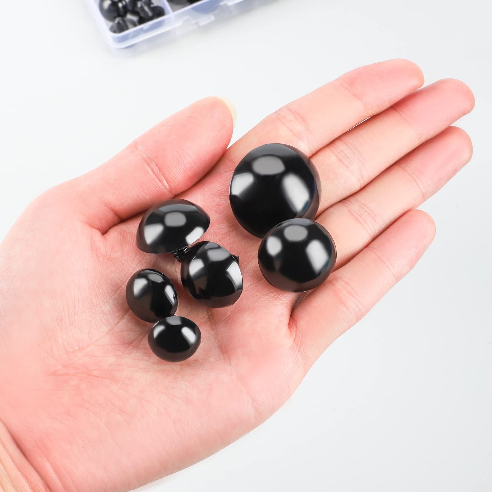 Hand holding various sizes of black safety eyes for crafts and stuffed animals. A clear plastic organizer with more eyes is visible in the background.