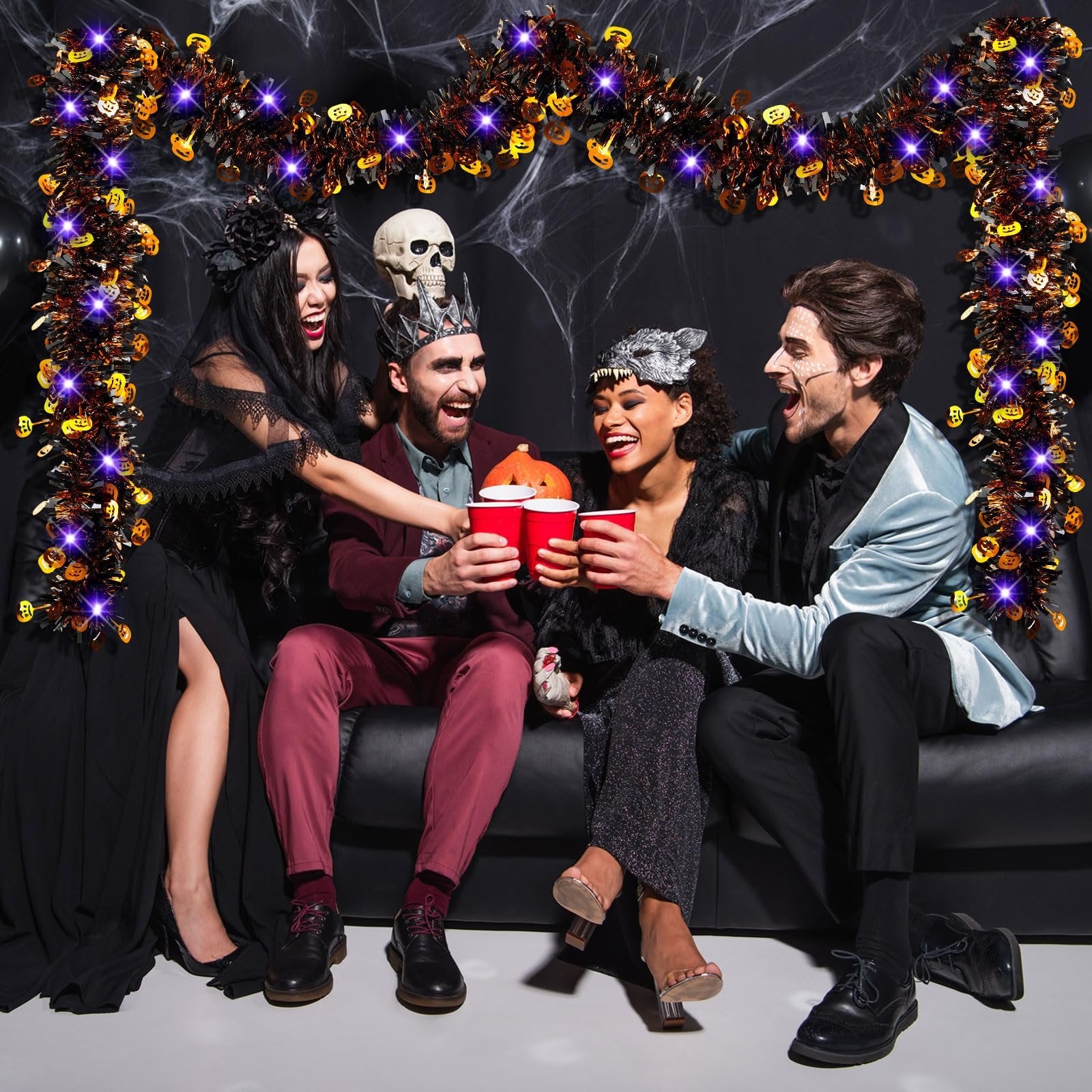 Four people in Halloween costumes toasting with red cups, with tinsel garland and spiderwebs in the background.