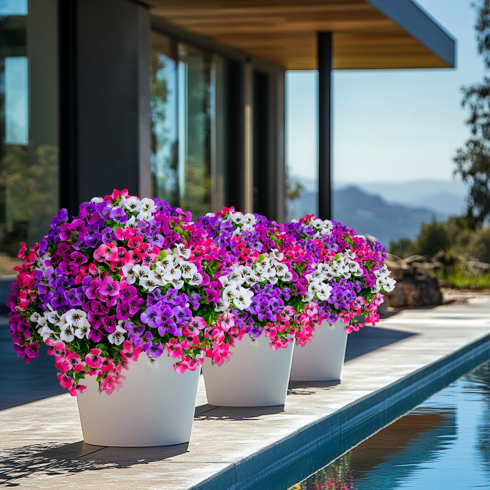 Three white planters filled with colorful artificial flowers arranged by a swimming pool with a modern house in the background.