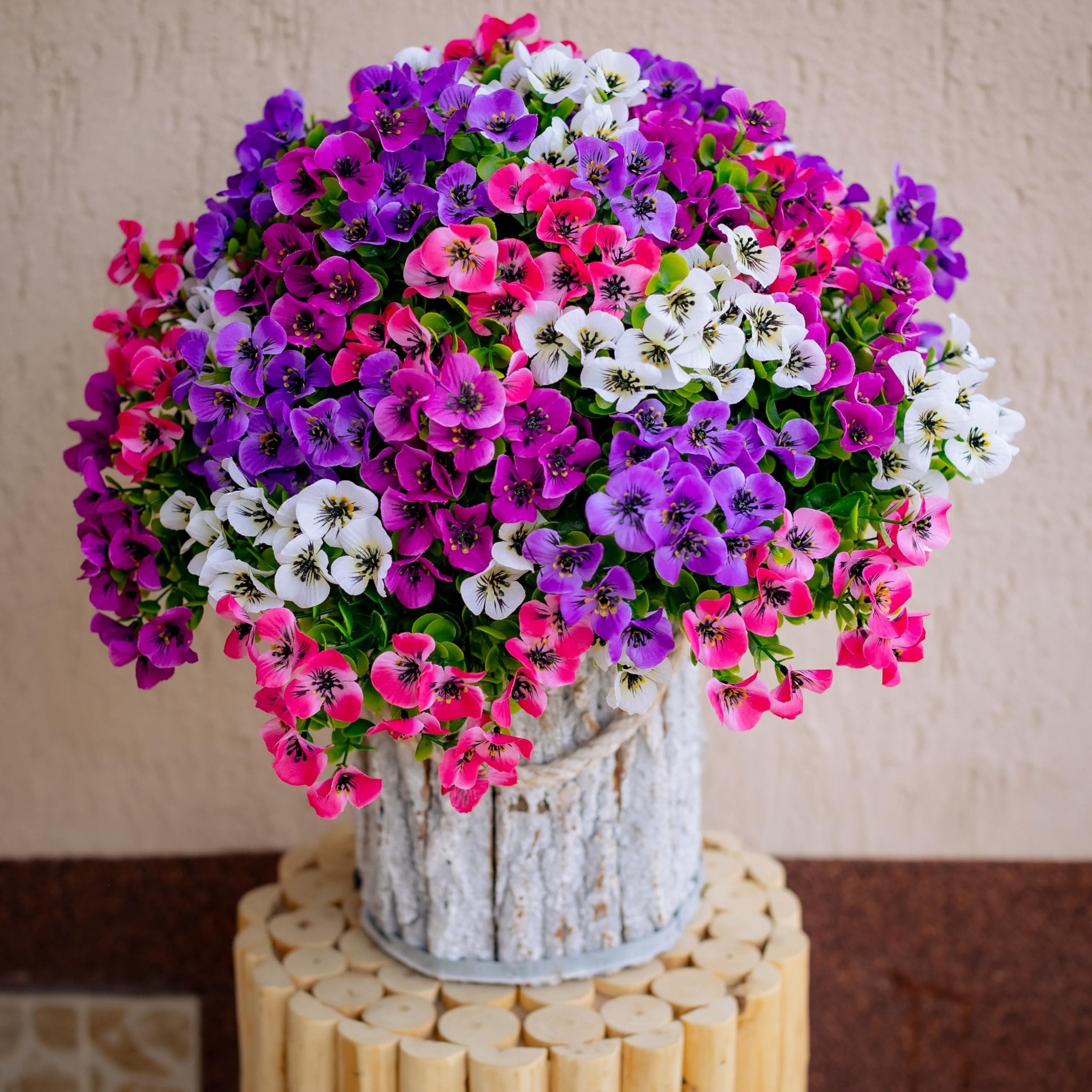 Bouquet of artificial flowers in pink, purple, and white, displayed in a rustic pot on a wooden stand.