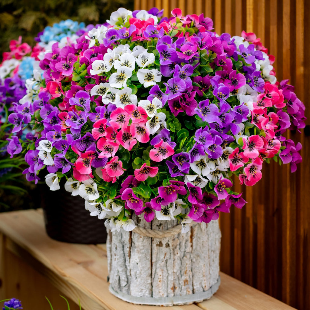 Close-up of a bouquet of artificial flowers in pink, purple, and white, with green leaves, in a textured pot on a wooden surface.