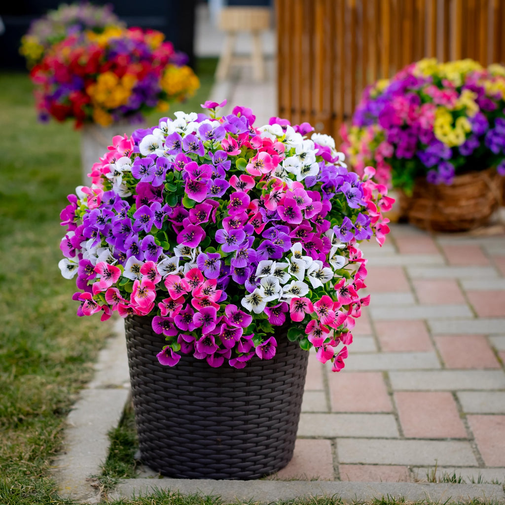 Artificial flowers in a dark brown woven pot on a paved walkway with grass and blurred background.