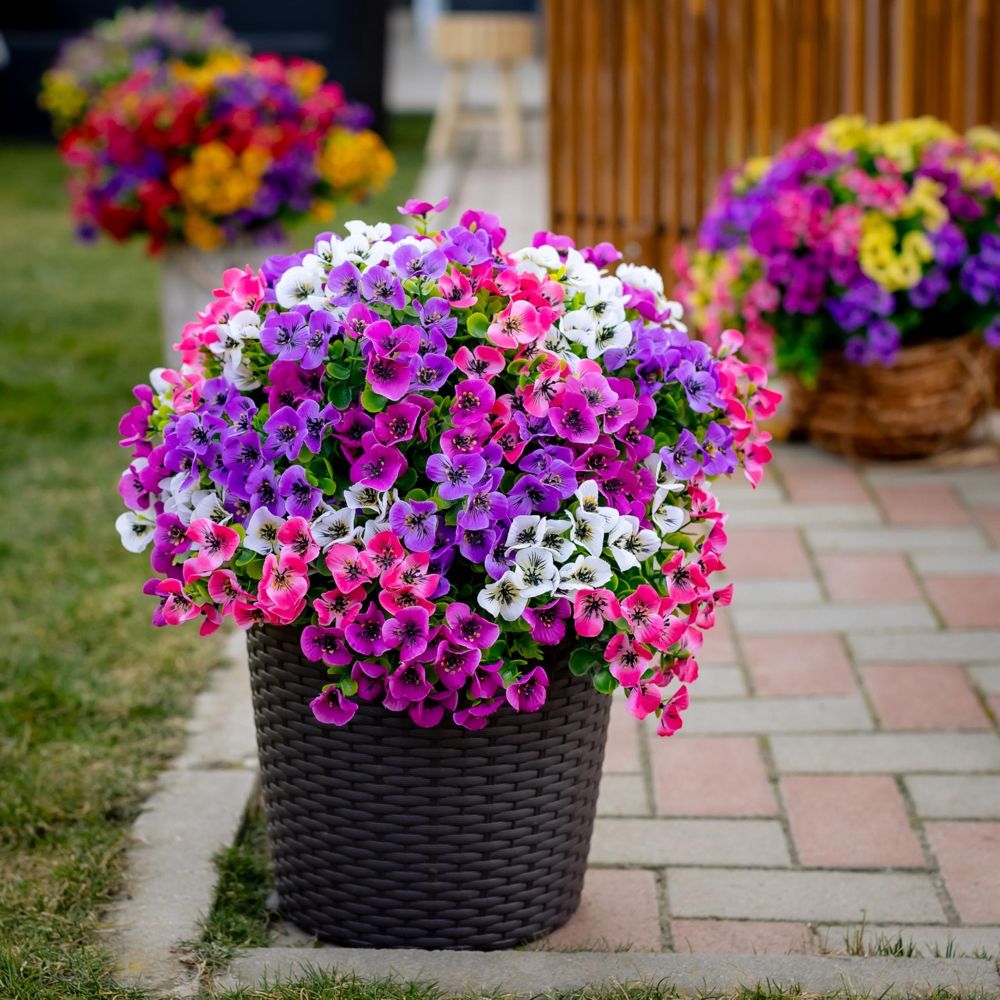 Artificial flowers in a dark brown woven pot on a paved walkway with grass and blurred background.