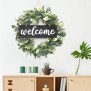 Artificial eucalyptus leaves wreath with a black welcome sign hanging on a white wall above a wooden cabinet.