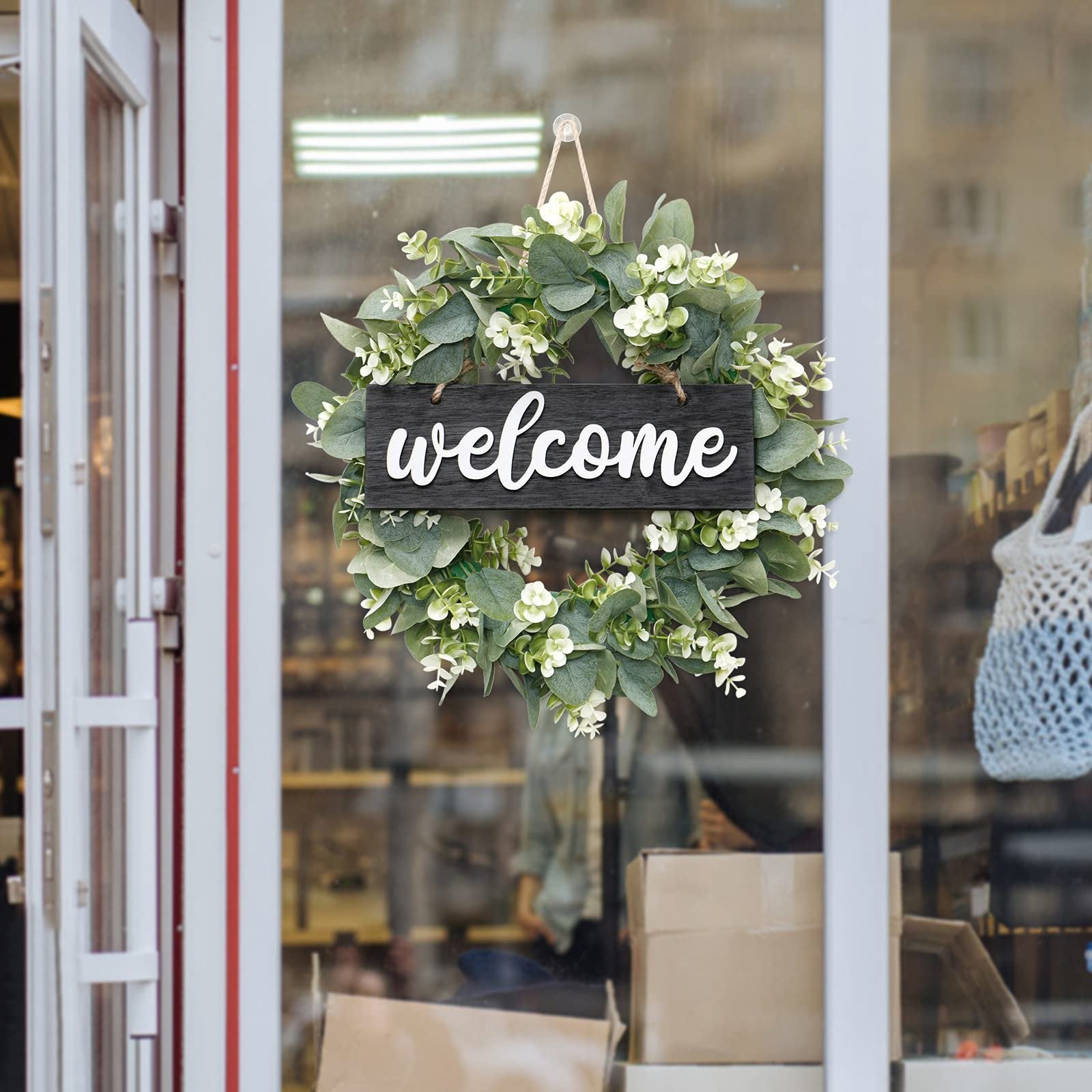 Artificial eucalyptus wreath with white flowers and a black welcome sign hanging on a glass door.