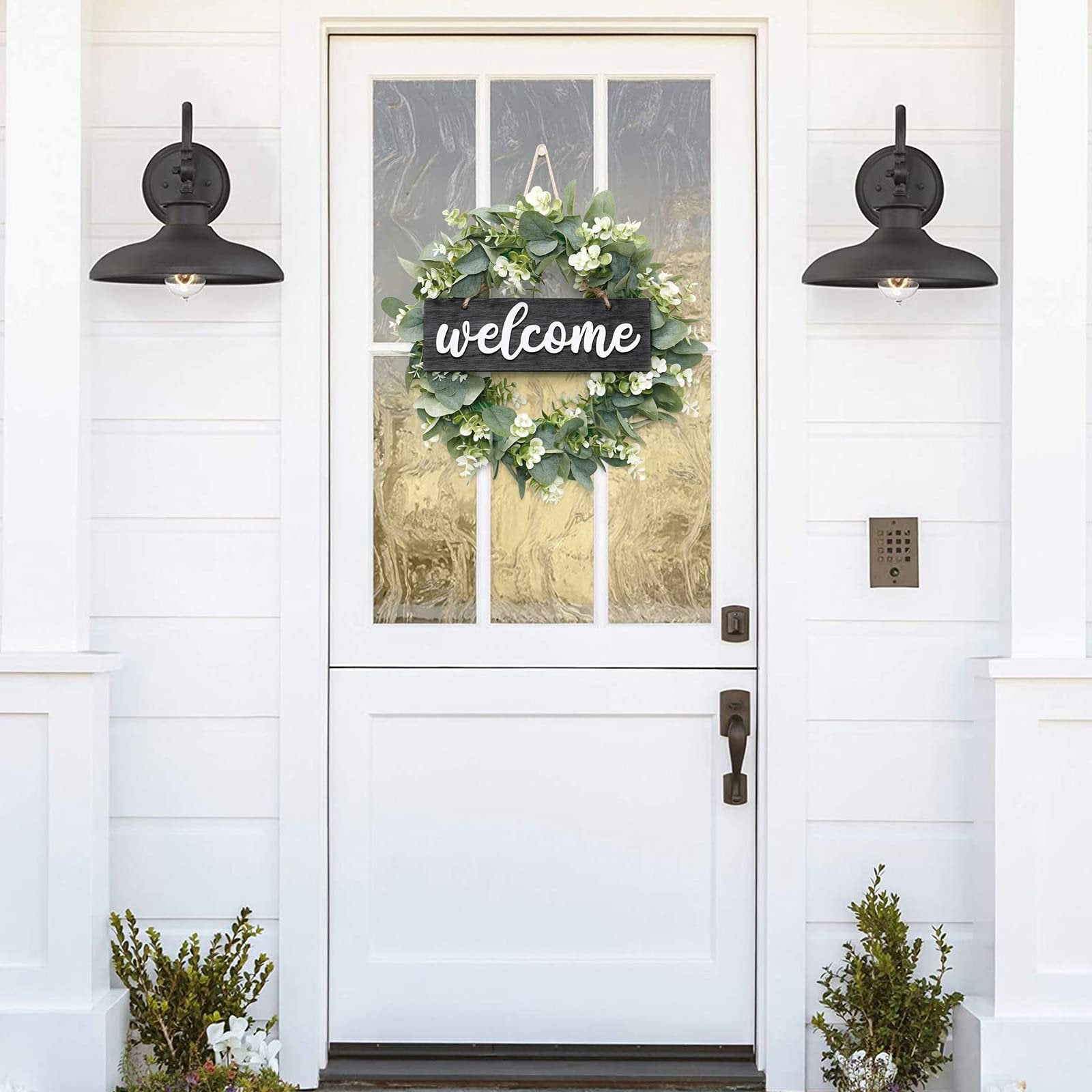 Artificial eucalyptus leaves wreath with a black welcome sign hanging on a white front door with wall sconces