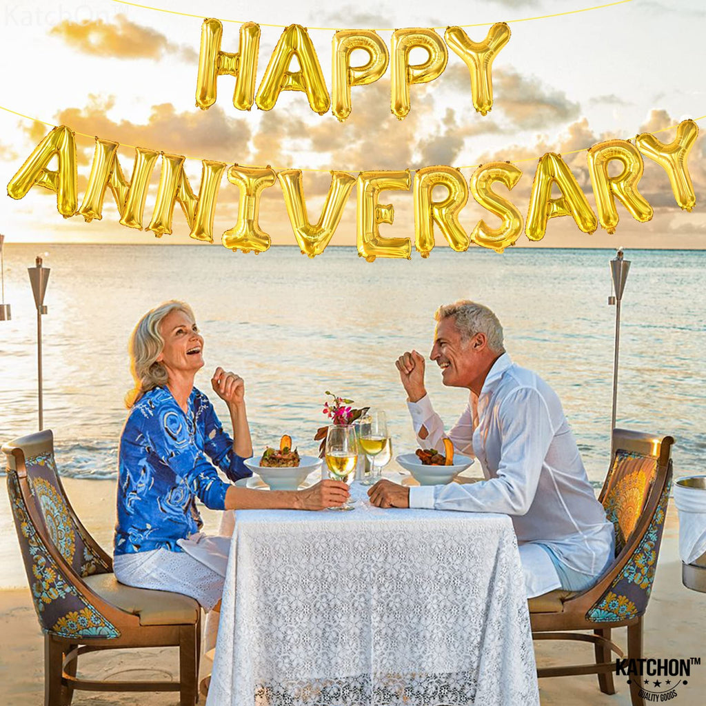 Couple celebrating anniversary at a beachside dinner with gold 'Happy Anniversary' balloons overhead
