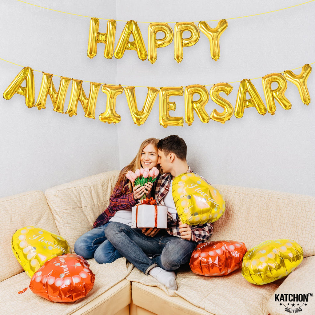 Couple celebrating anniversary with gold letter balloons and heart-shaped balloons on a couch.
