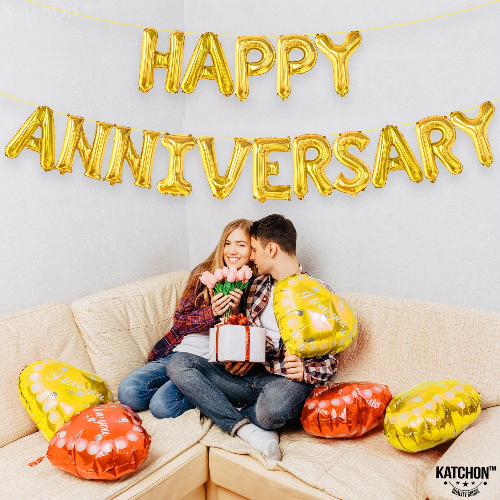Couple celebrating anniversary with gold letter balloons and heart-shaped balloons on a couch.