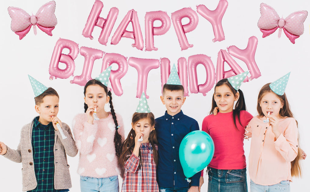Children wearing party hats celebrate with pink foil 'Happy Birthday' balloons and bow balloons.