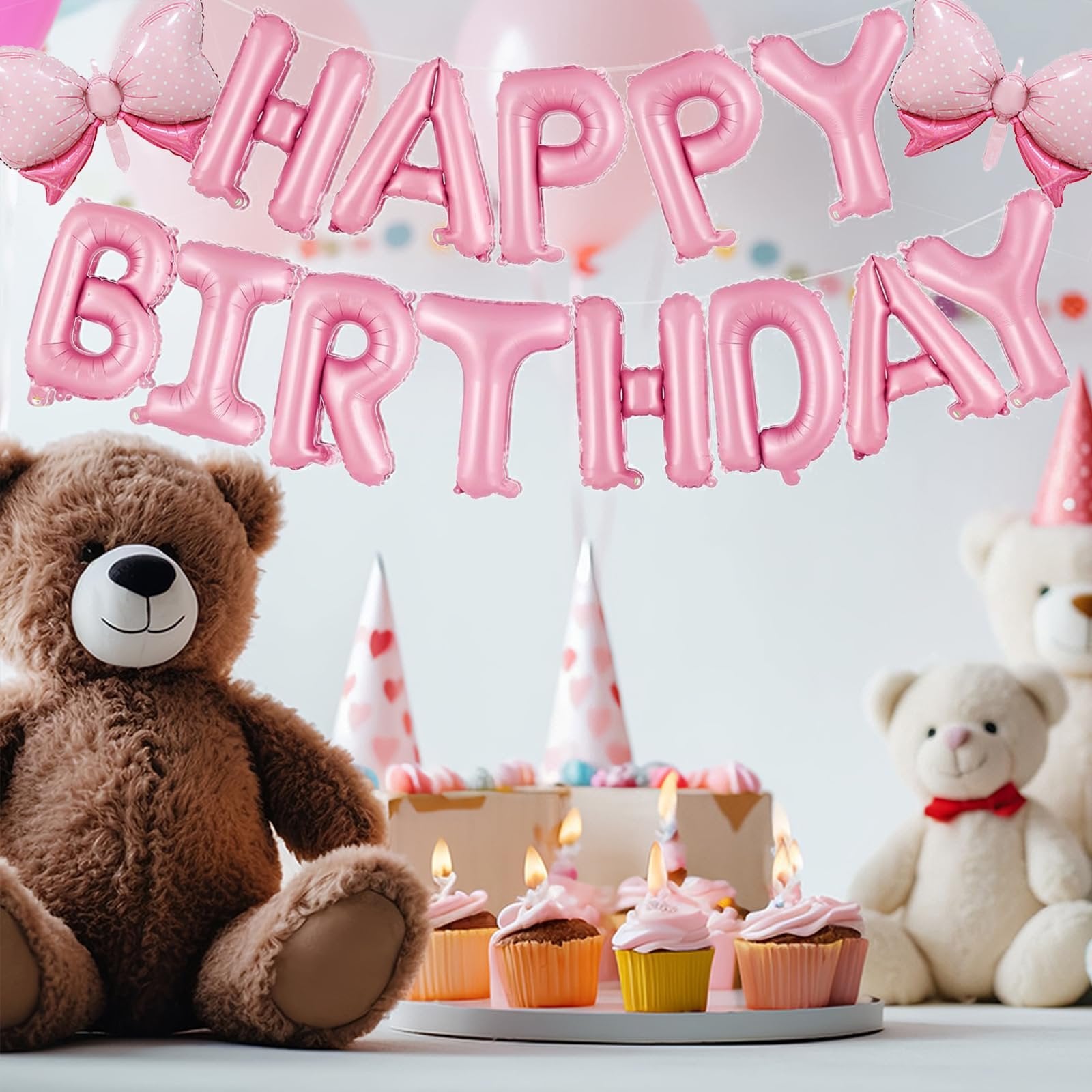 Pink foil 'Happy Birthday' balloon banner with two bow balloons, set up for a party with teddy bears and cupcakes.