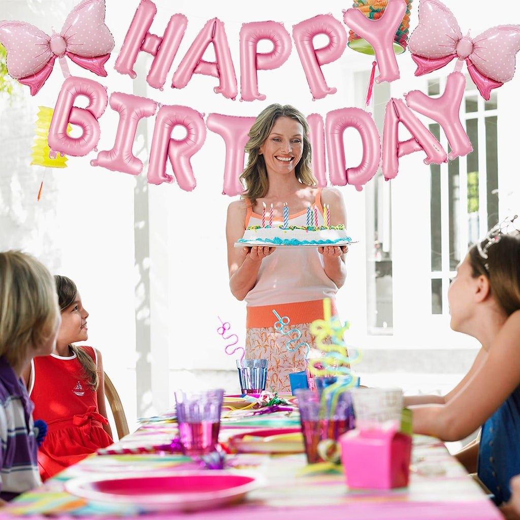Woman holding birthday cake with pink foil balloons spelling 'HAPPY BIRTHDAY' behind her at a party