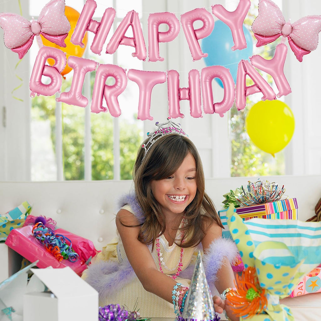 Girl wearing a tiara and boa smiles while opening presents, with pink foil 'Happy Birthday' balloons overhead.
