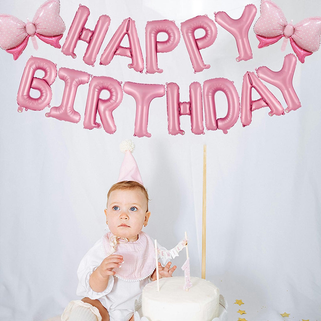 Baby girl in pink party hat sits by a cake with pink 'Happy Birthday' balloons overhead