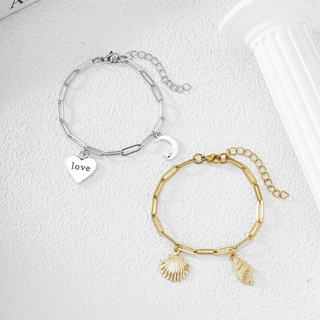 Two paperclip chain bracelets, one silver with heart and moon charms, one gold with shell charms, on a textured white surface.