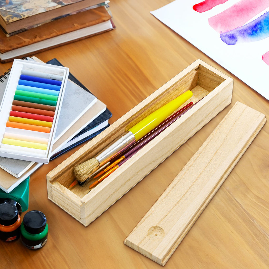 Unfinished pine wood box with sliding lid open, showing paint brushes and a yellow marker inside, next to a box of pastels and art supplies.