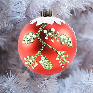 Red shatterproof Christmas ornament with green glitter snowflake design and white glitter accents, hanging on a white artificial tree.