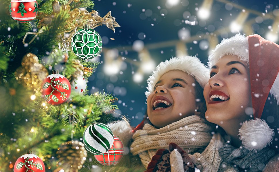 Mother and child in Santa hats looking up at a decorated Christmas tree with ornaments and falling snow.