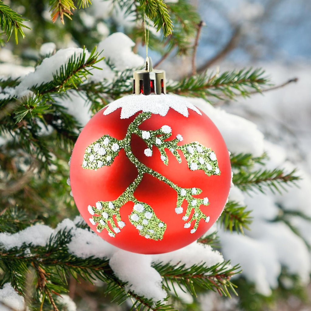 Red Christmas ornament with green glittery branch design hanging on a snow-covered pine tree branch