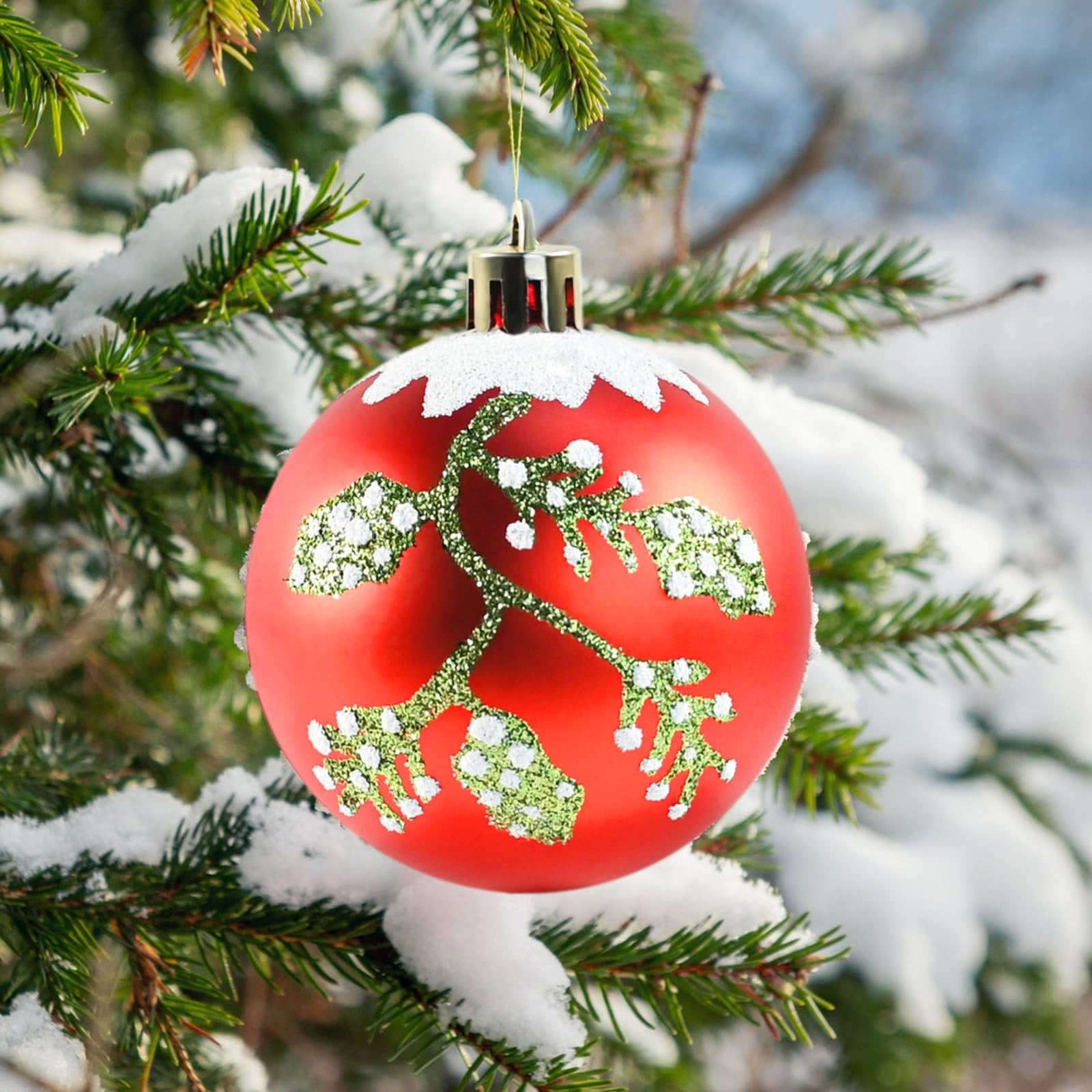 Red Christmas ornament with green glittery branch design hanging on a snow-covered pine tree branch