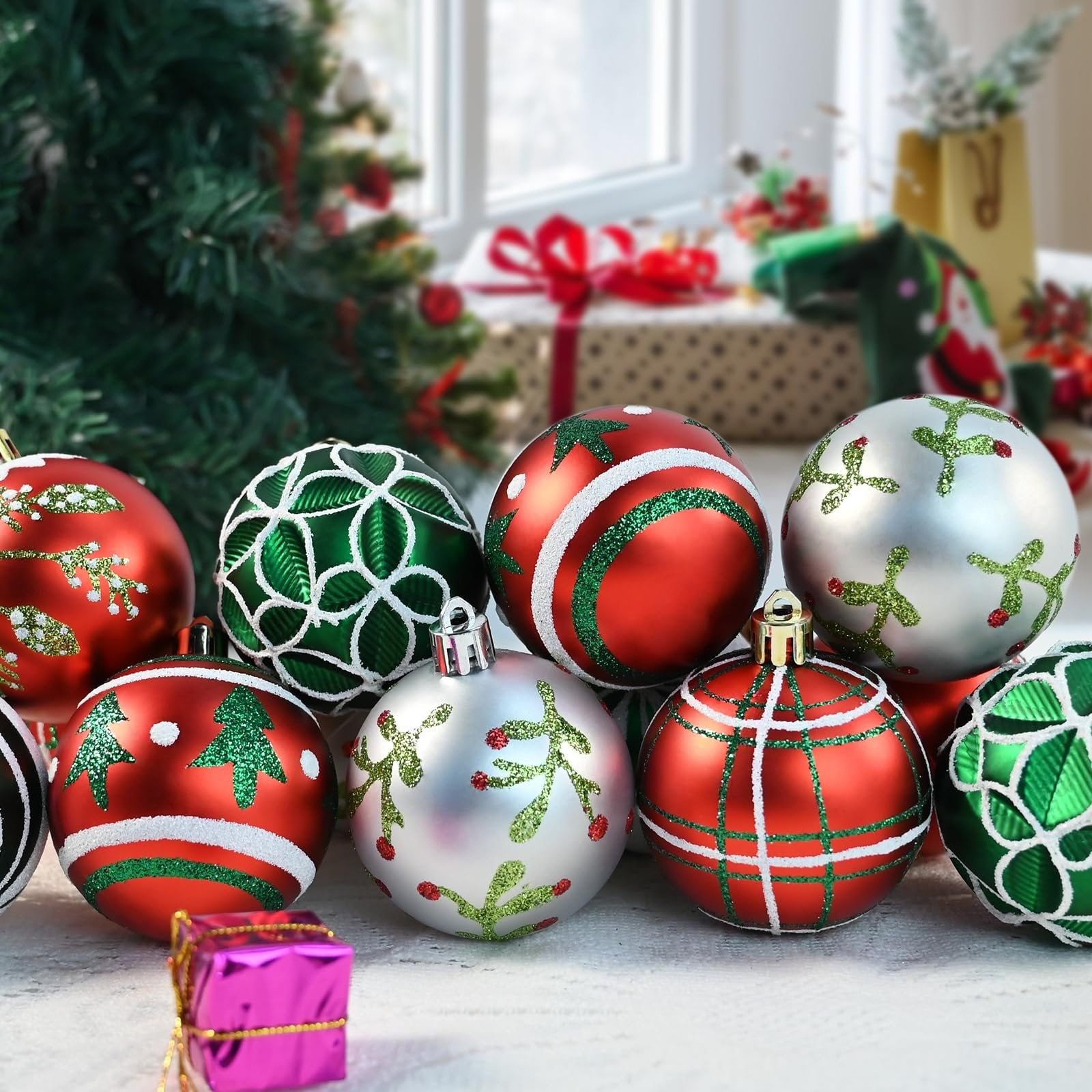 Assortment of red, green, and silver shatterproof Christmas ball ornaments with glitter designs, arranged in front of a decorated Christmas tree and gifts.