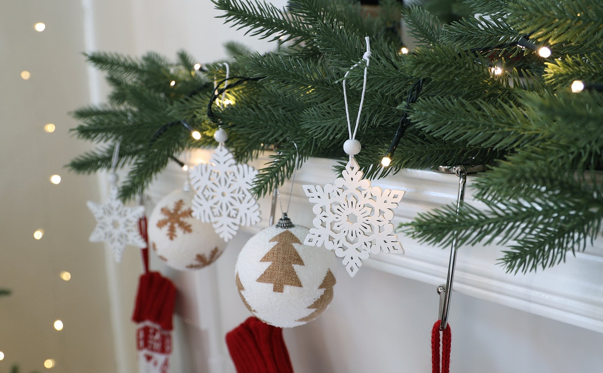Close-up of white wooden snowflake ornaments and fabric ball ornaments with tree designs hanging from a Christmas tree branch with lights.