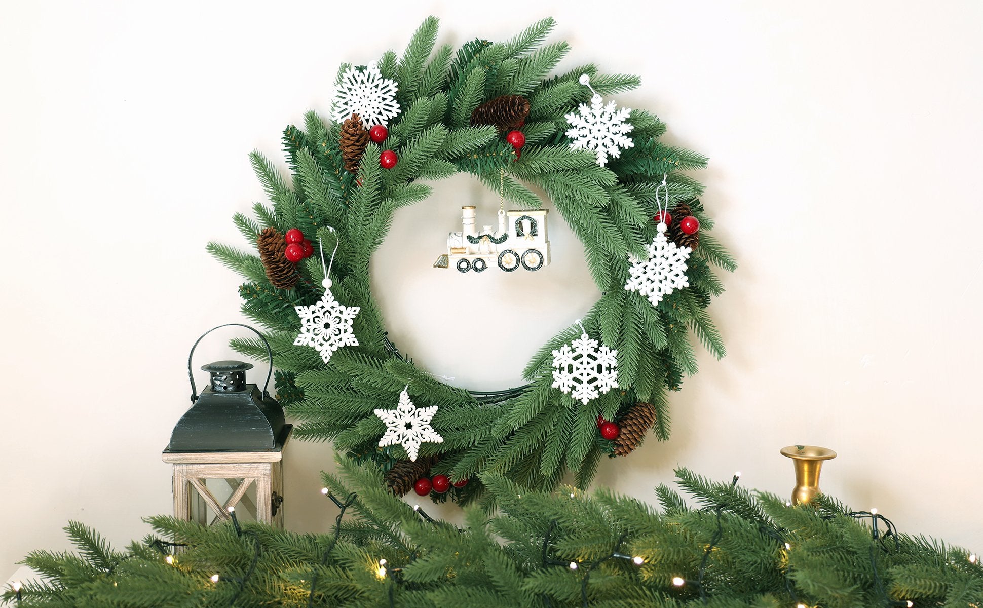 Christmas wreath with white snowflake ornaments and a toy train decoration, next to a lantern and string lights.