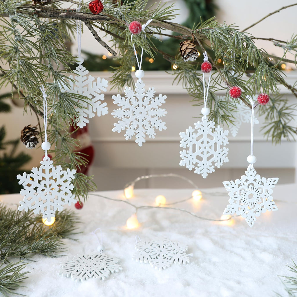 White wooden snowflake ornaments hanging from a pine branch with red berries and pinecones, set against a snowy background with fairy lights.