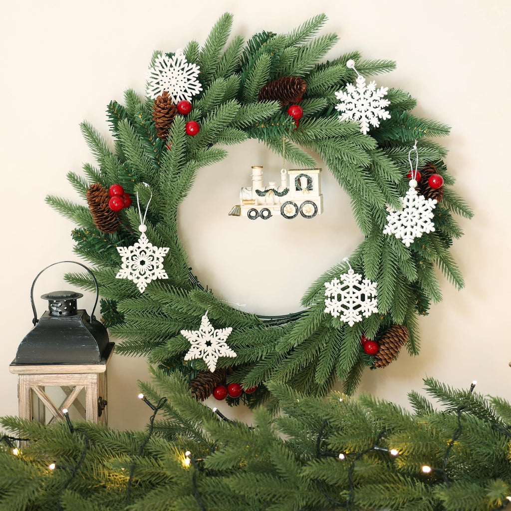 Christmas wreath with snowflake ornaments, pine cones, red berries, and a train decoration, next to a lantern and string lights.