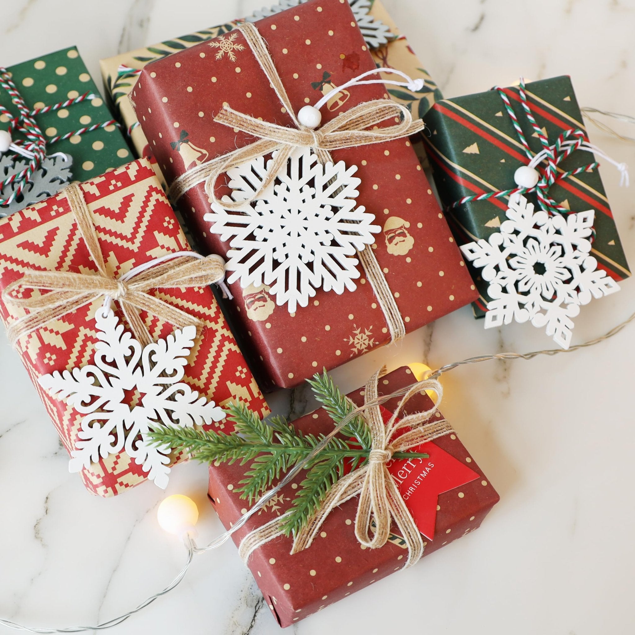 Several Christmas gifts wrapped in various festive papers, adorned with white wooden snowflakes and twine, with fairy lights.