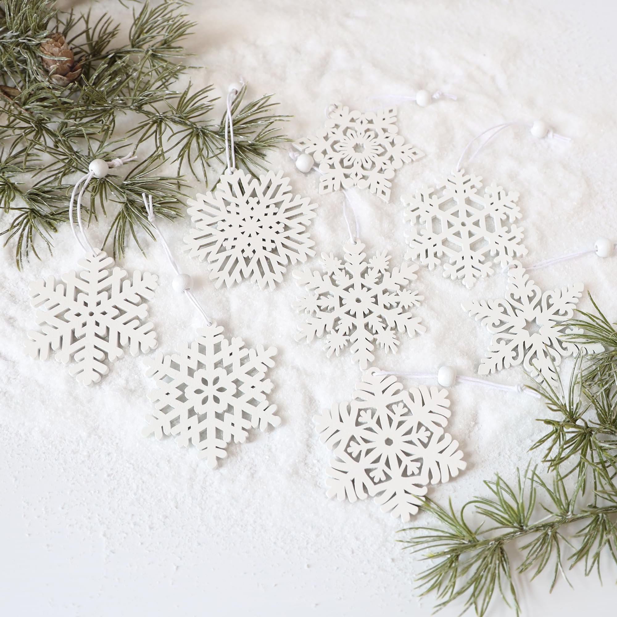 White wooden snowflake ornaments scattered on faux snow with pine branches and a pinecone.