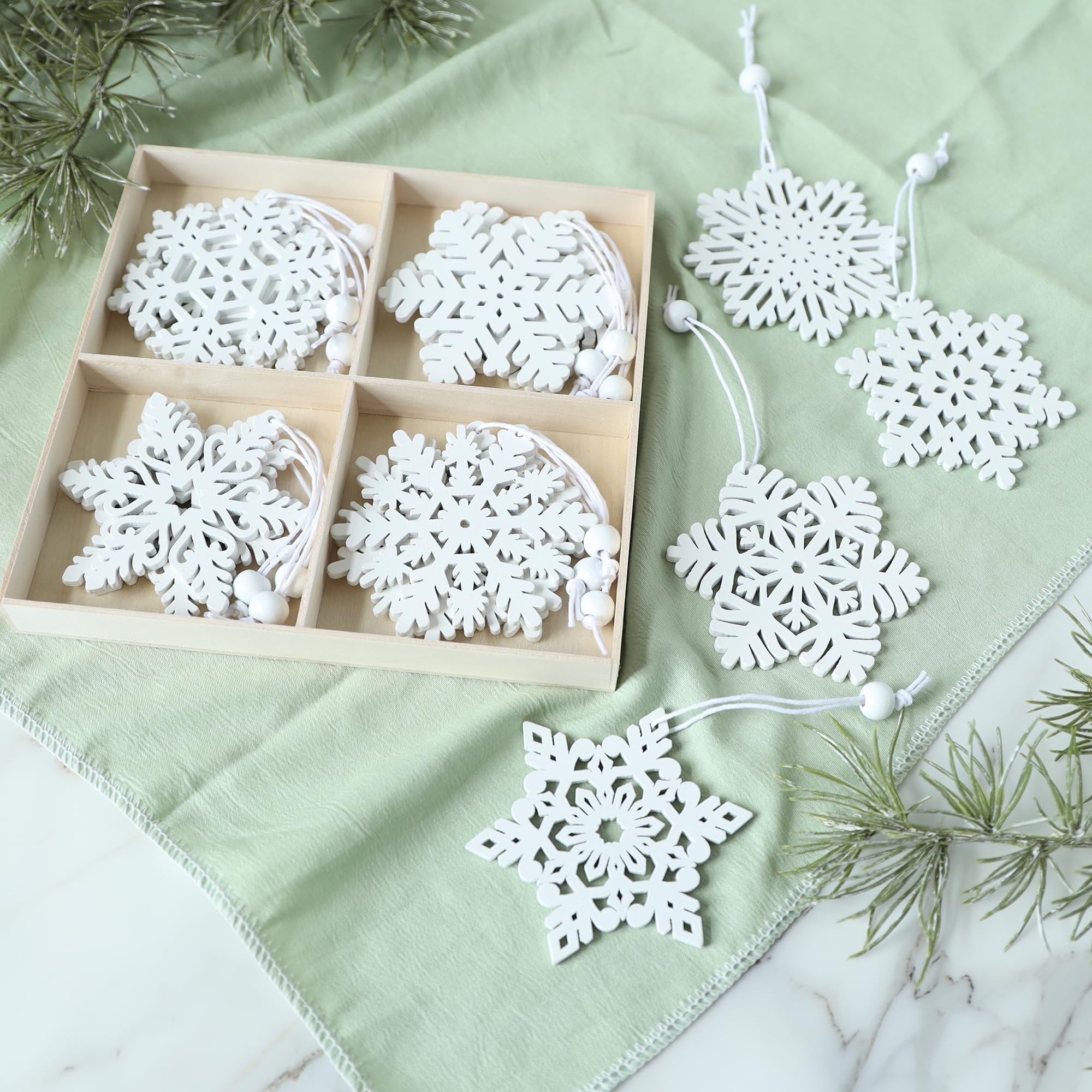 Set of white wooden snowflake ornaments in a wooden box and scattered on a green cloth with pine branches.