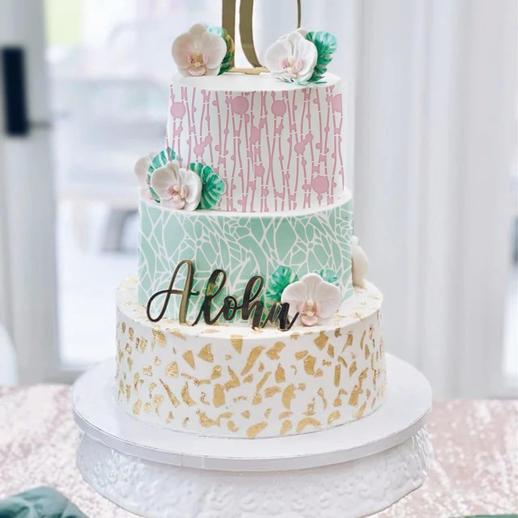 Three-tier decorated cake with floral accents and 'Aloha' topper, displayed on a white cake stand.