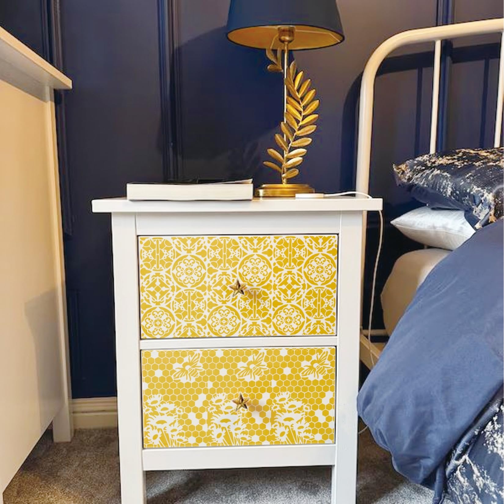 White nightstand with two drawers decorated with yellow and white floral and honeycomb stencils, next to a bed.