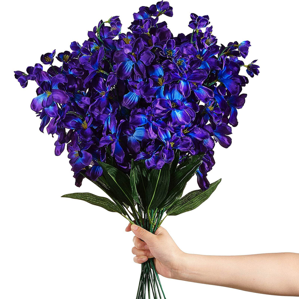 Hand holding a bouquet of 16 artificial blue-purple orchid stems with green leaves against a white background.