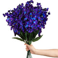 Hand holding a bouquet of 16 artificial blue-purple orchid stems with green leaves against a white background.