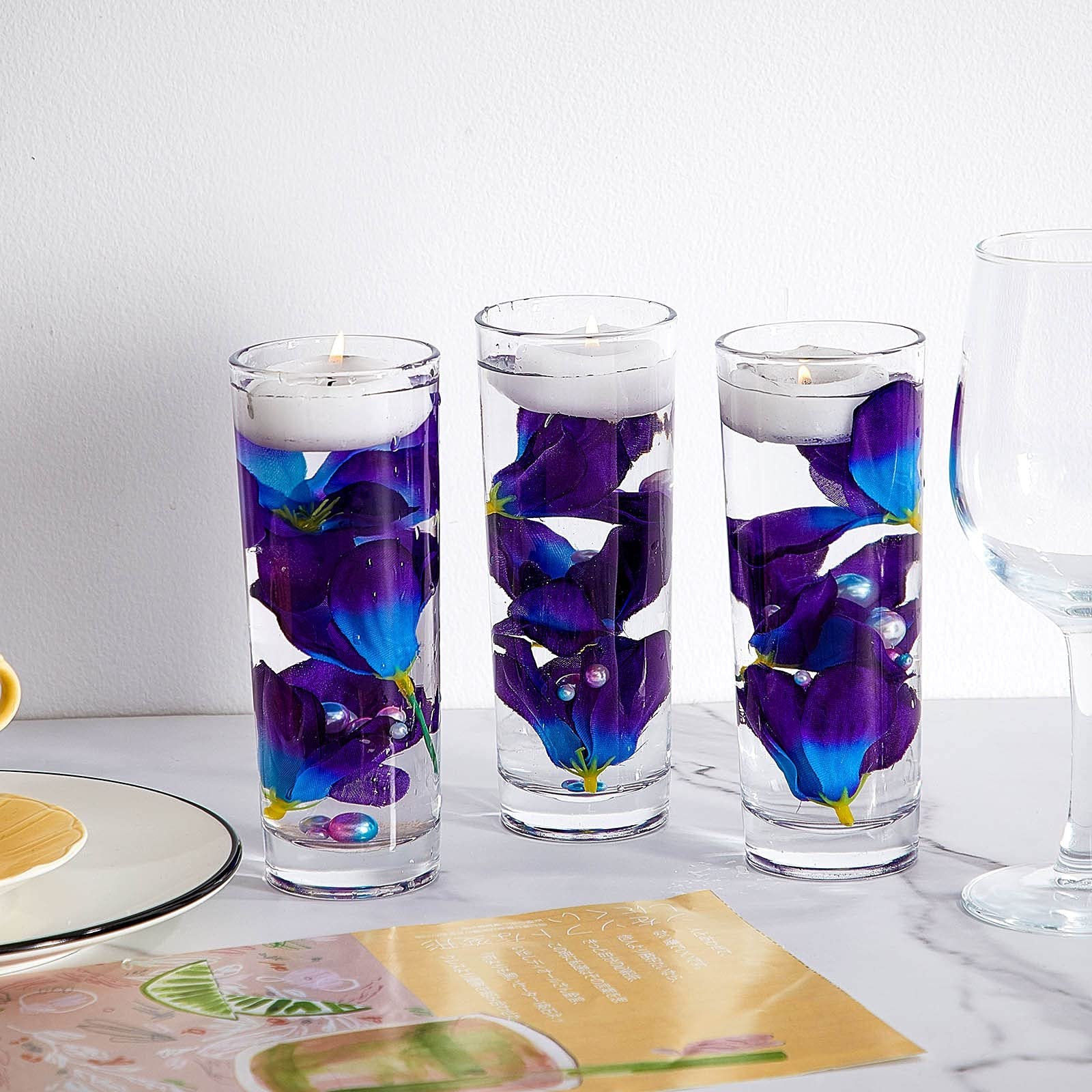 Three clear glass cylinders filled with water, blue-purple artificial orchid petals, and floating candles.