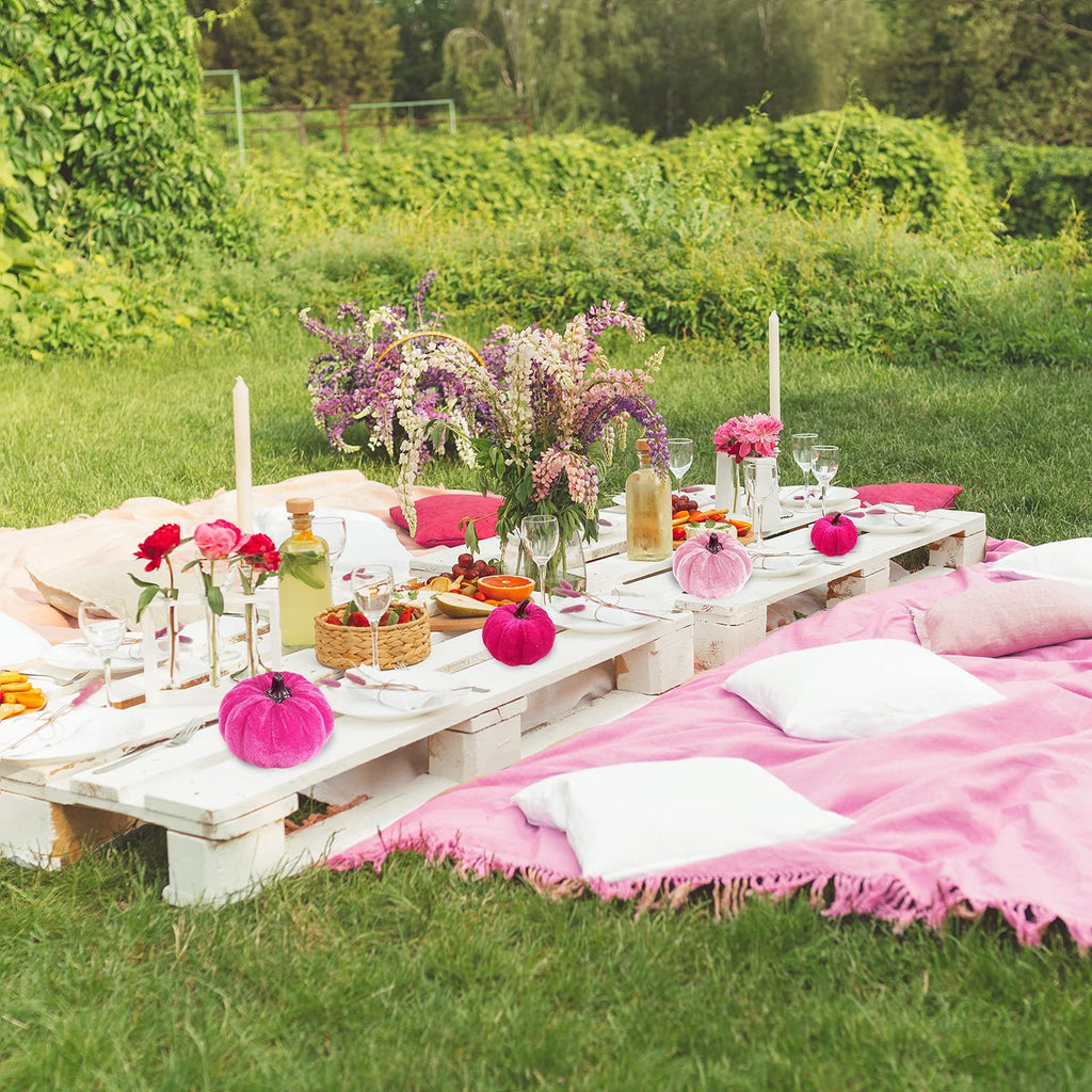 Outdoor picnic table set with pink velvet pumpkins, flowers, and food on a sunny day.