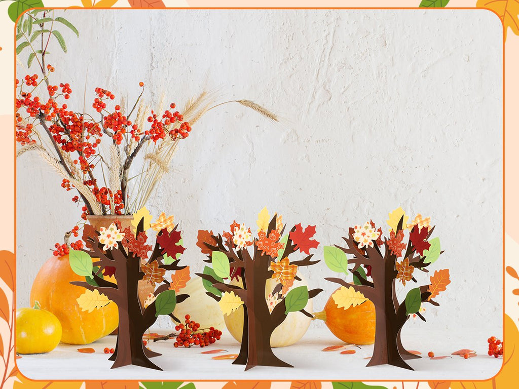 Three cardboard thankful trees decorated with fall leaves and flowers, set against a white wall with pumpkins and autumn foliage.