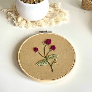 Embroidery hoop with tan fabric and a stitched floral design featuring purple flowers and green leaves.