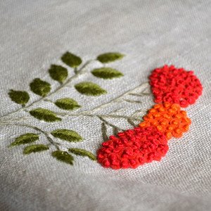 Close-up of embroidered floral branch with red and orange flowers on linen fabric.