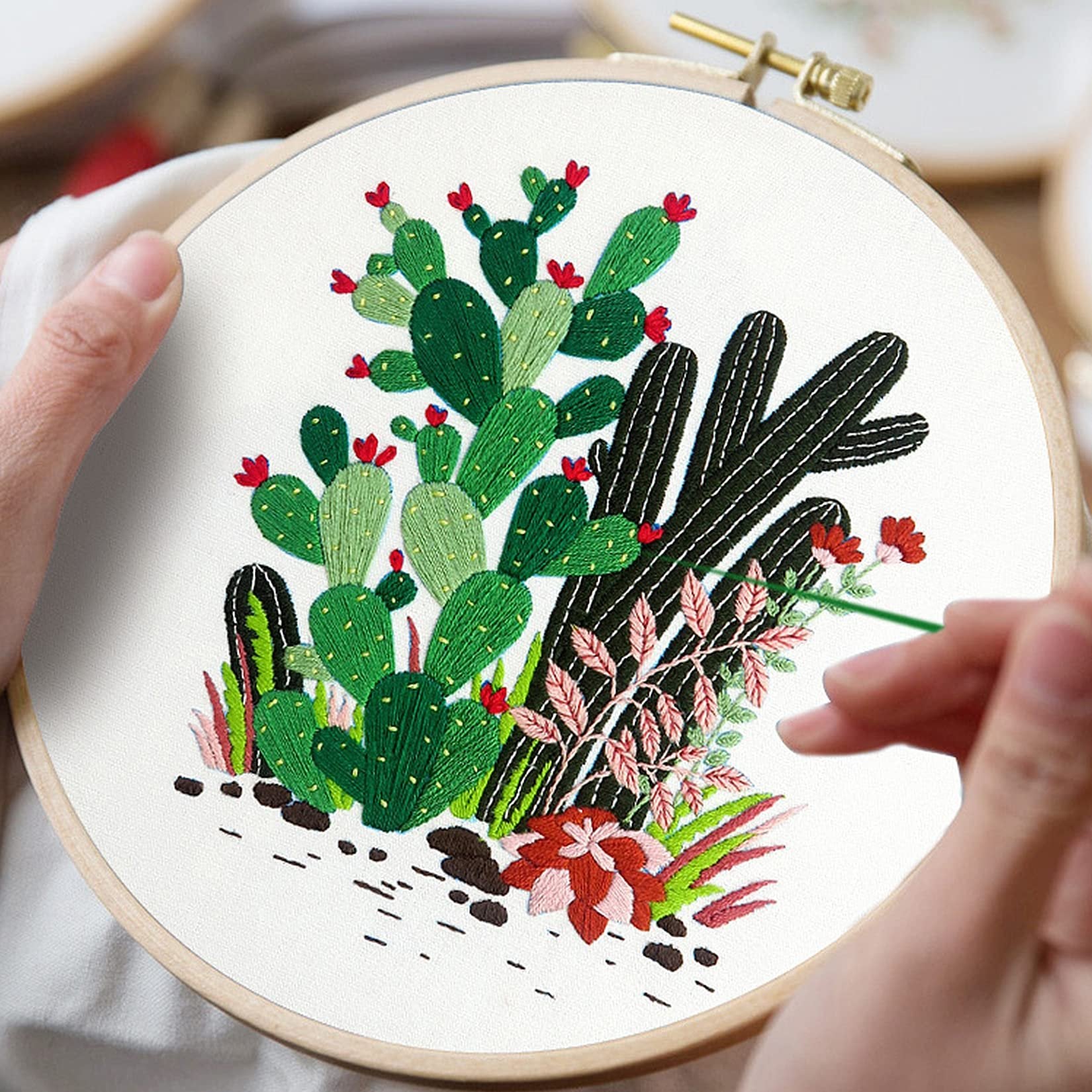 Embroidery hoop with a cactus and succulent design being stitched by hands, close-up view.