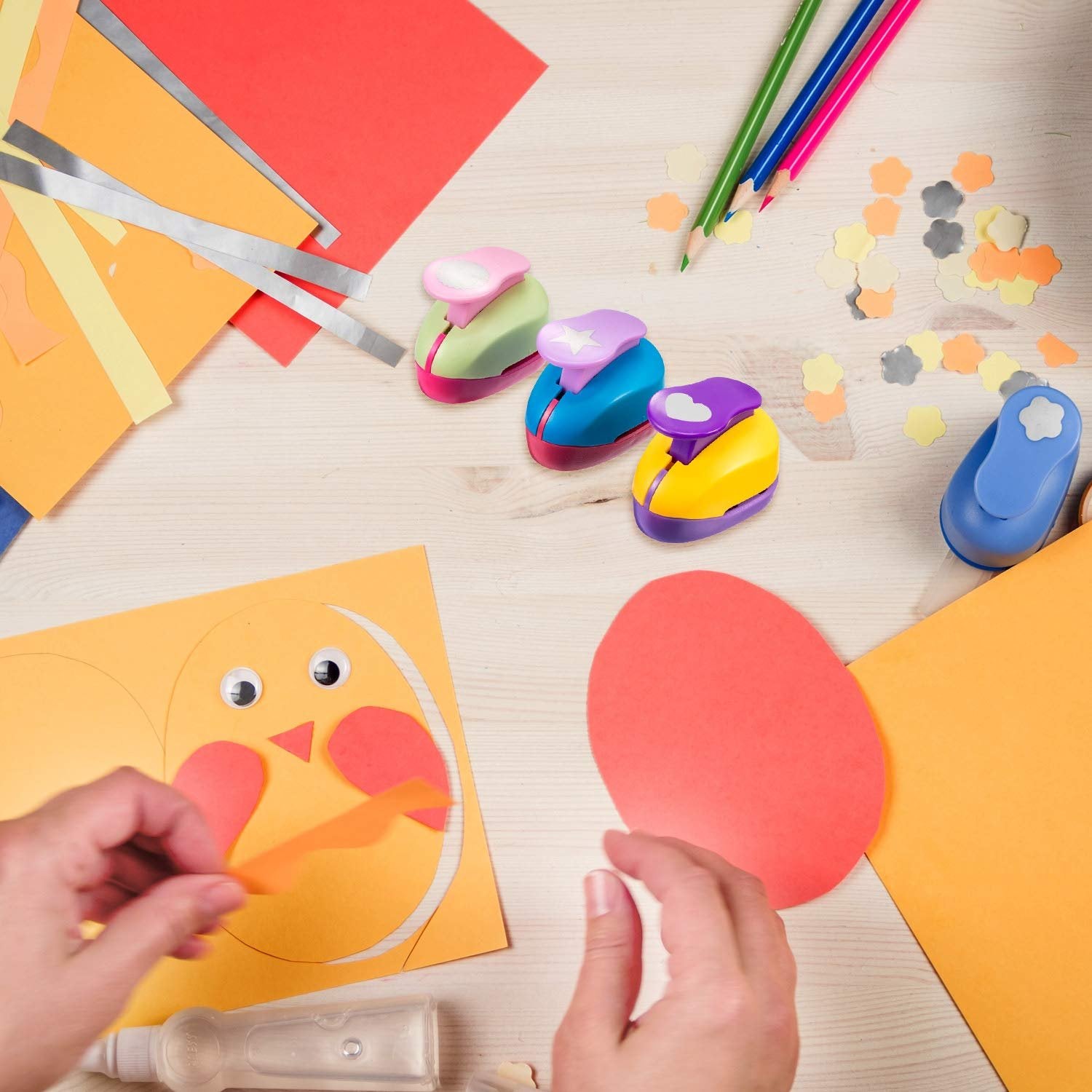Hands crafting a chick from paper, surrounded by craft punches, colored paper, pencils, and confetti shapes.