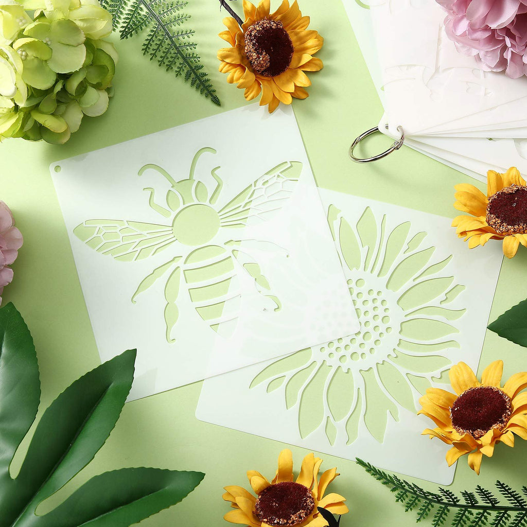 Set of reusable painting stencils with bee and sunflower designs, held together by a metal ring, surrounded by artificial flowers and greenery.