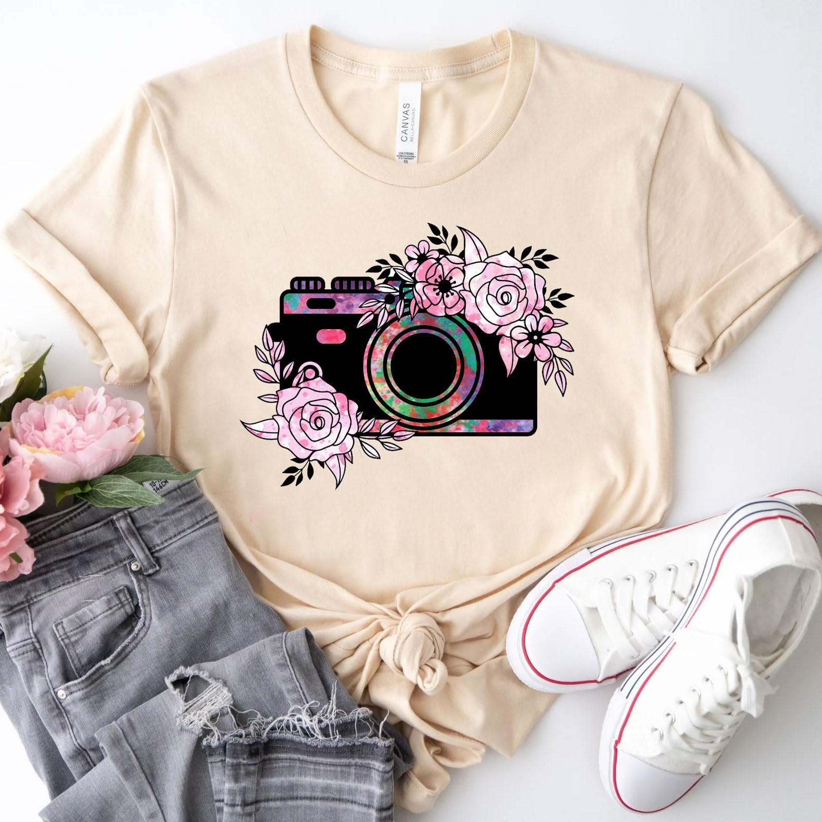 Beige t-shirt with a black camera graphic surrounded by pink flowers, styled with gray jeans and white sneakers.
