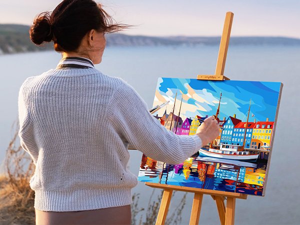 Woman painting a colorful harbor scene on canvas outdoors, with a body of water and hills in the background.