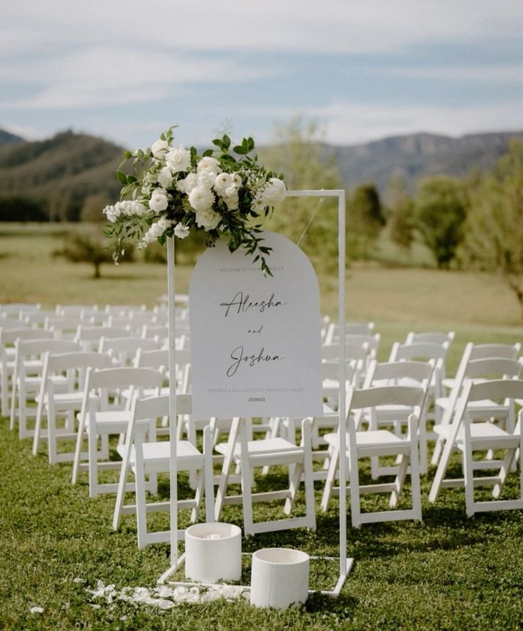 Wedding ceremony setup with rows of white chairs and a welcome sign decorated with white flowers.