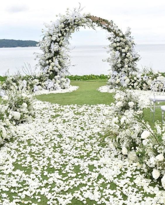 Wedding ceremony aisle decorated with white flower petals and an archway adorned with white flowers, overlooking the ocean.