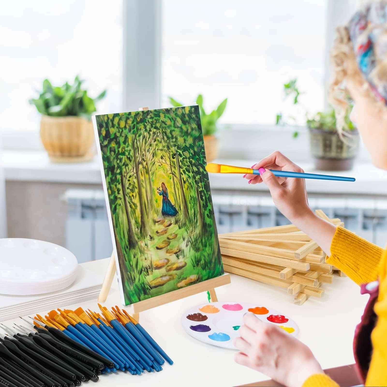 Woman painting a forest scene on canvas with brushes and palette, surrounded by art supplies